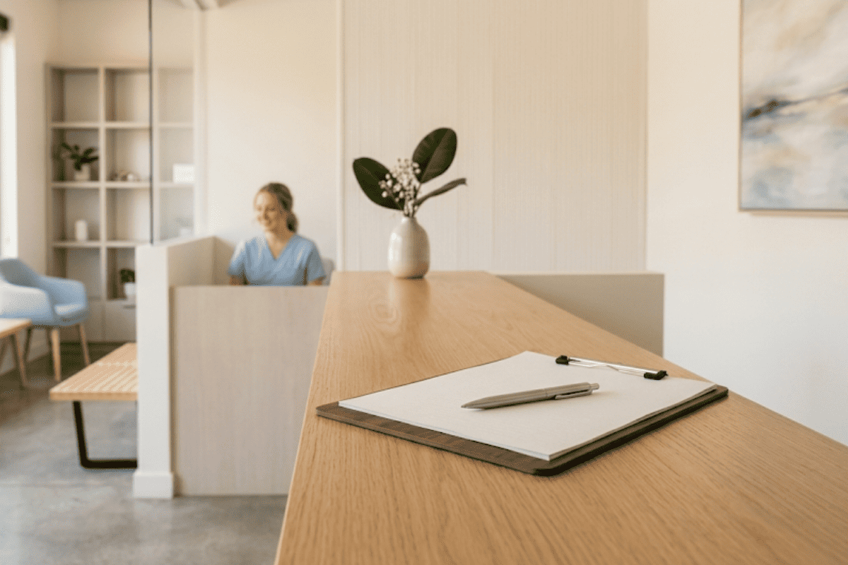cant-afford-to-get-my-tooth-pulled - Dentistry For You OKC | OKC Dentist Reception desk in a bright lobby; a receptionist sits behind a partition, with a clipboard and pen on the foreground counter.