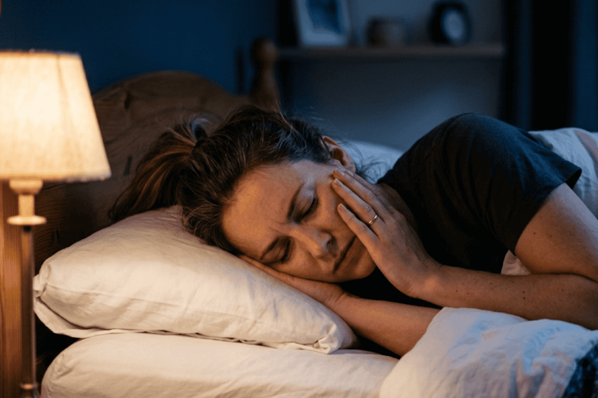 Woman resting on a bed at night, hand to her face, eyes closed, illuminated by a warm bedside lamp.